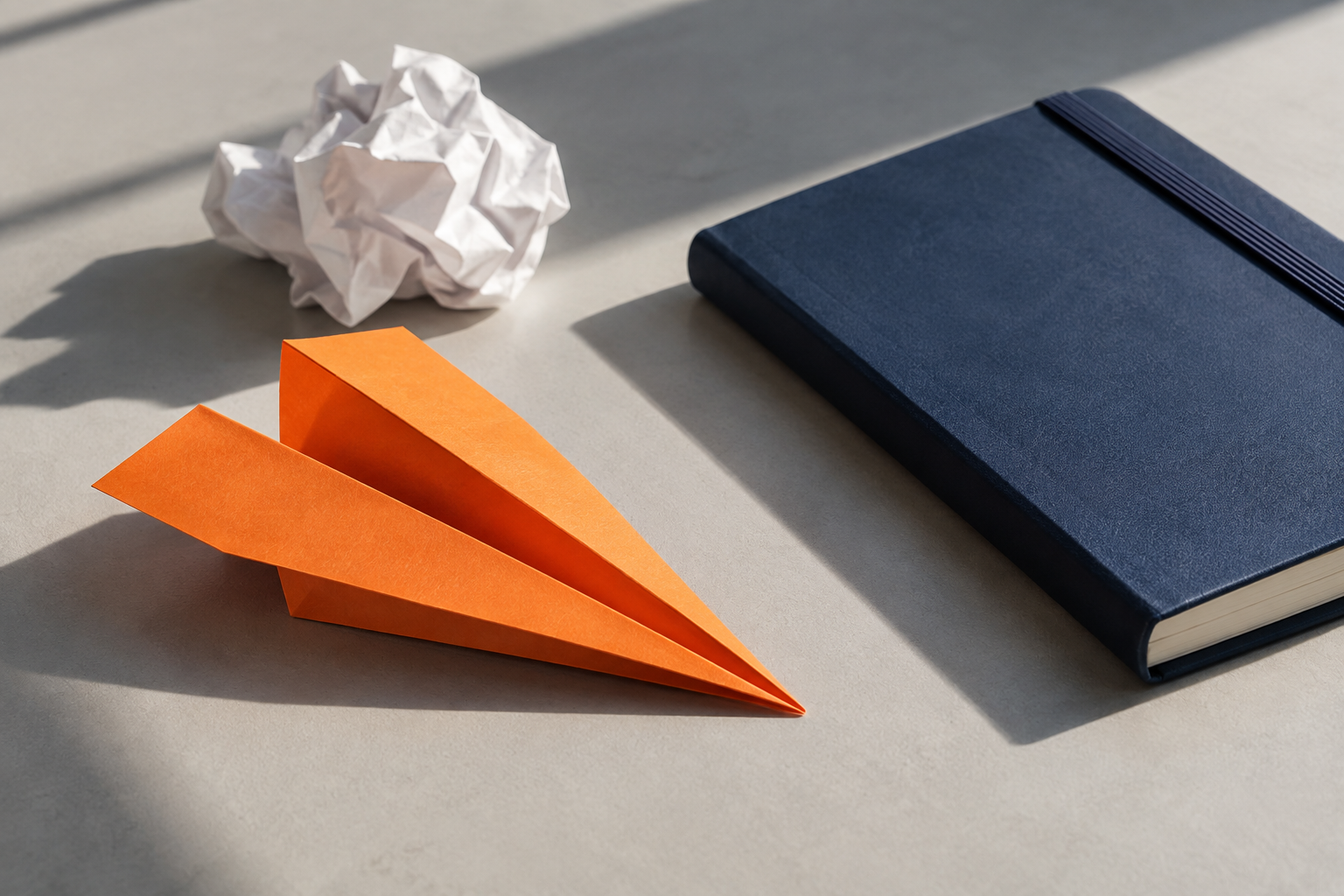 Avião de papel laranja ao lado de um caderno azul e de uma folha amassada.